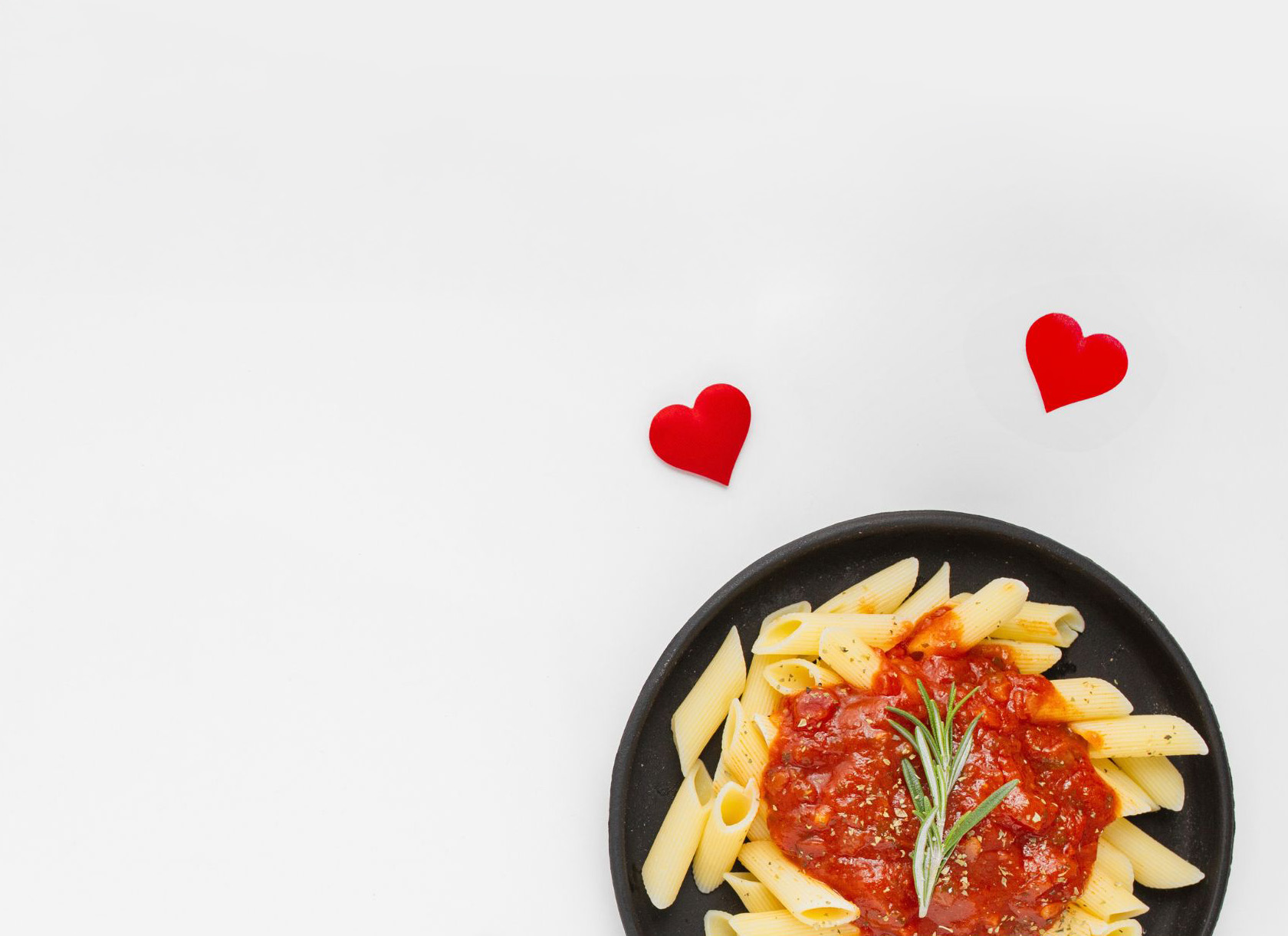 A plate of penne with tomato sauce and rosemary, with two red fabric hearts beside it