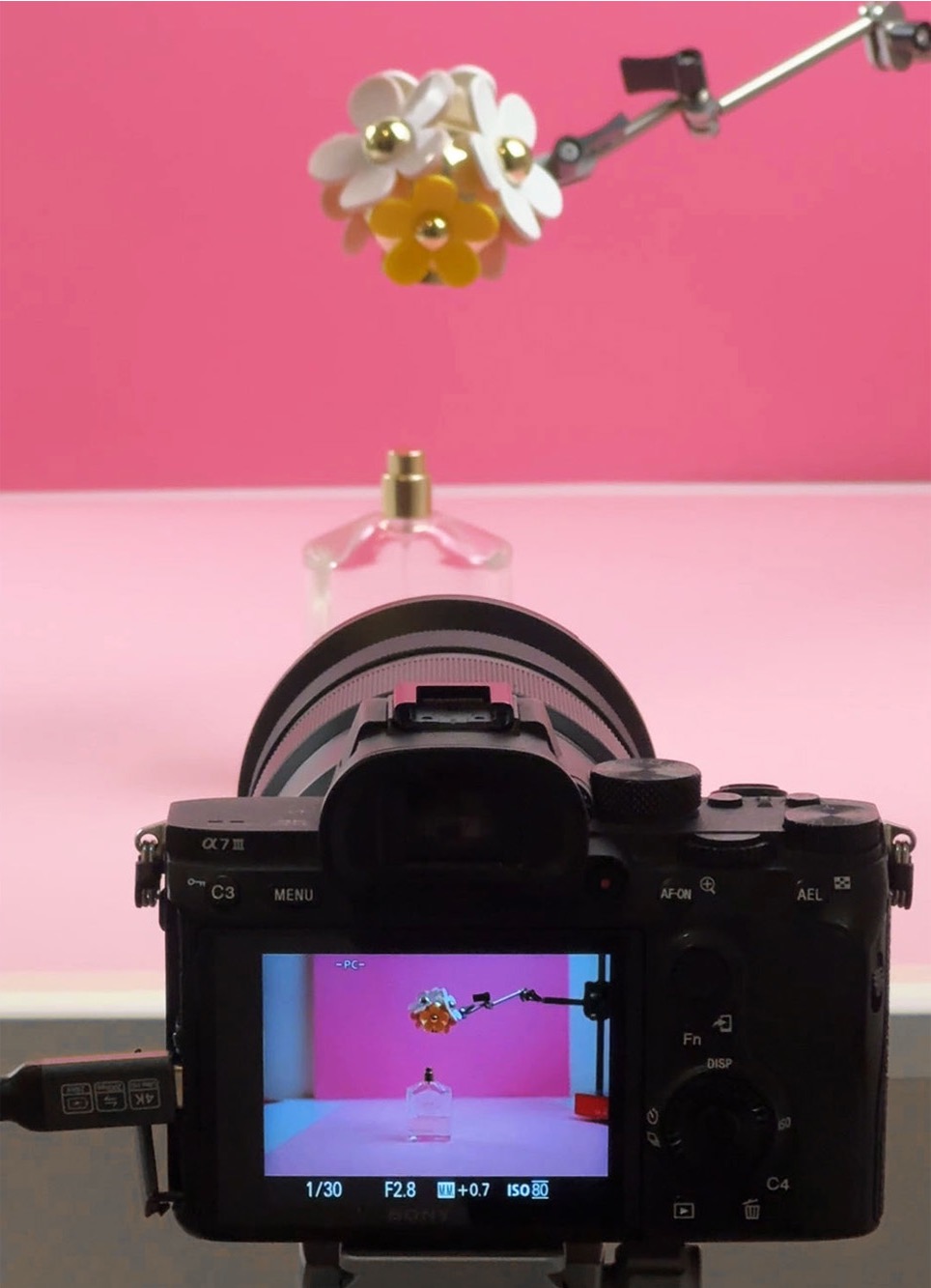 Behind the scenes: a camera filming a perfume bottle and flowers held on a rig in front of a pink background